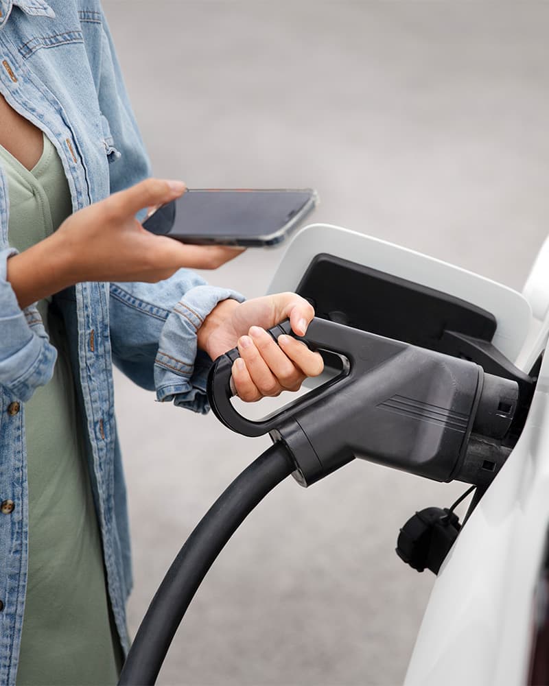 Laden und gleichzeitig verbunden – Elektromobilität im Alltag Frau hält Smartphone und lädt gleichzeitig ein Elektroauto an einer Schnellladestation